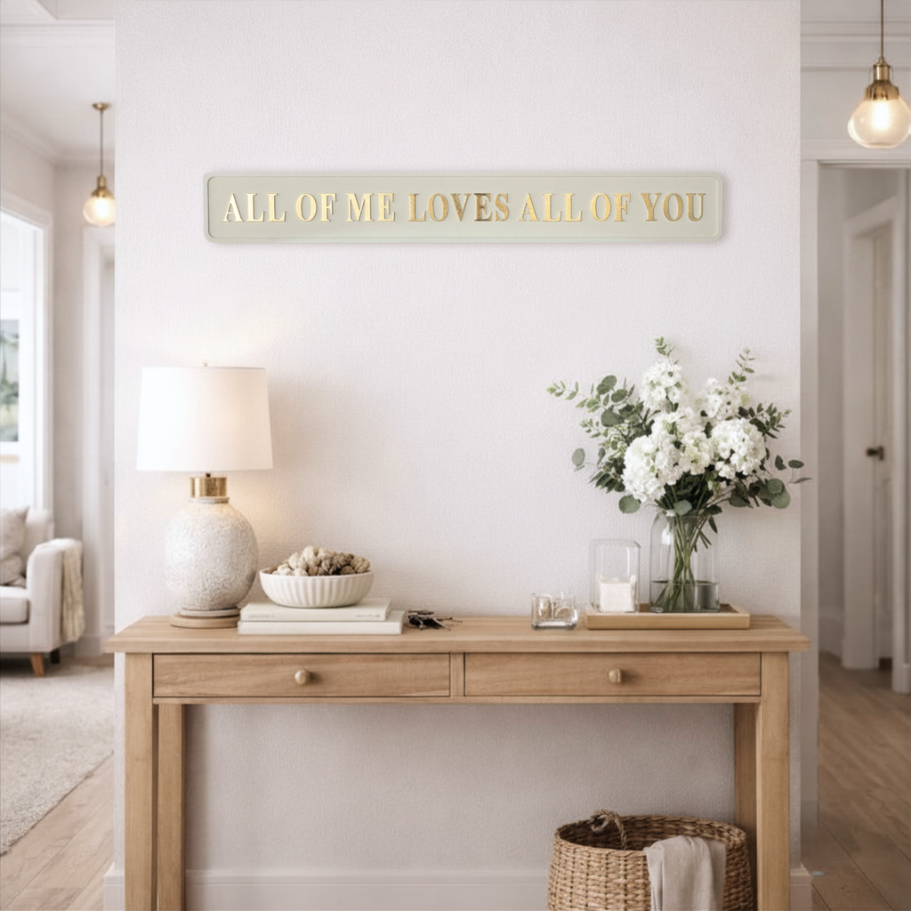 Cream and gold statement sign saying ALL OF ME LOVES ALL OF YOU above a wooden console table with decorative items in a bright hallway.