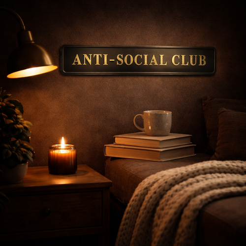 Cosy room with 'Anti-Social Club' sign in black and gold, lit candle, books, and mug.