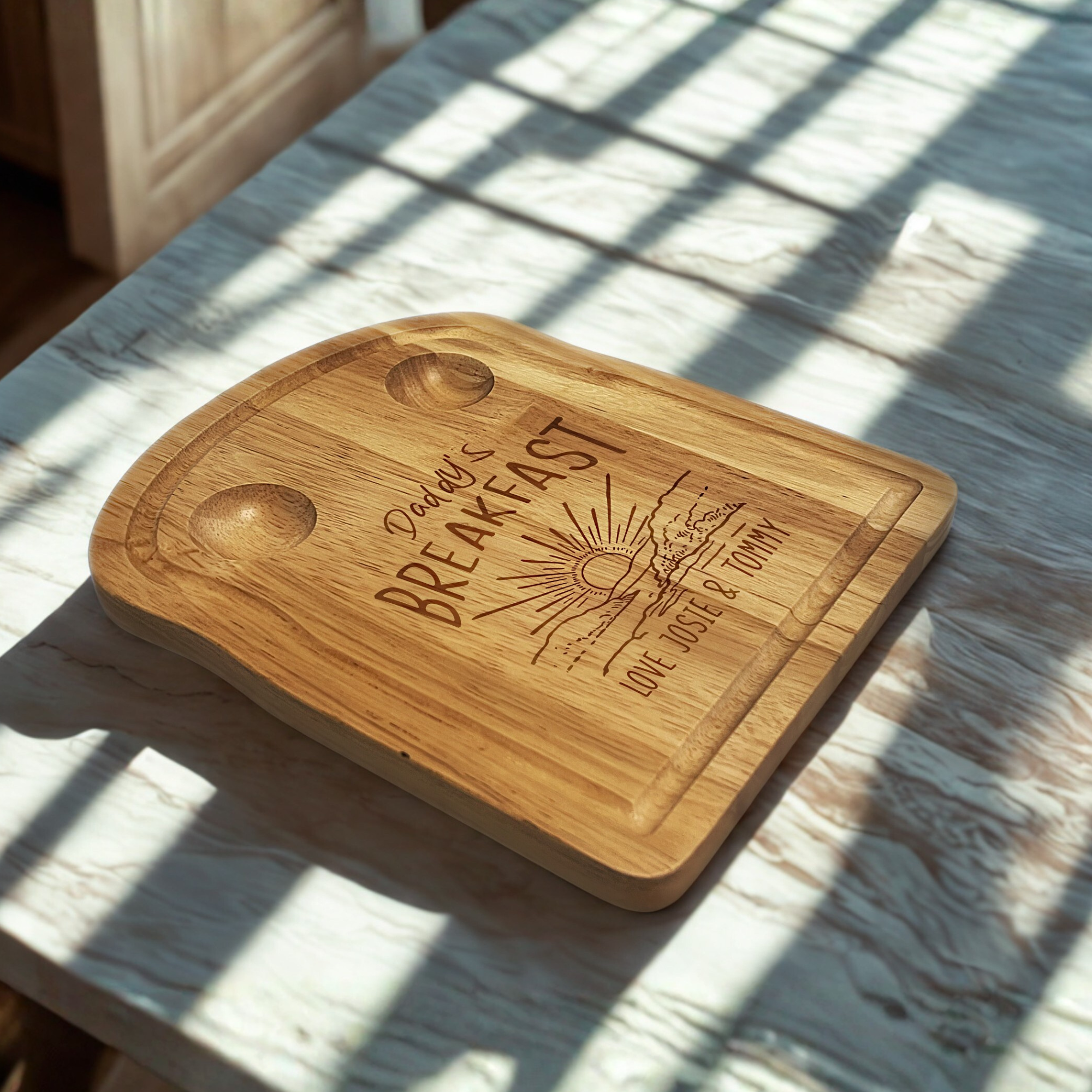 Wooden toast shaped serving board with 2 carved egg holders in the top, an engraved illustration of a sunset with 'Daddy's Breakfast' above it and a personalised message engraved at the bottom. On a sunny kitchen counter