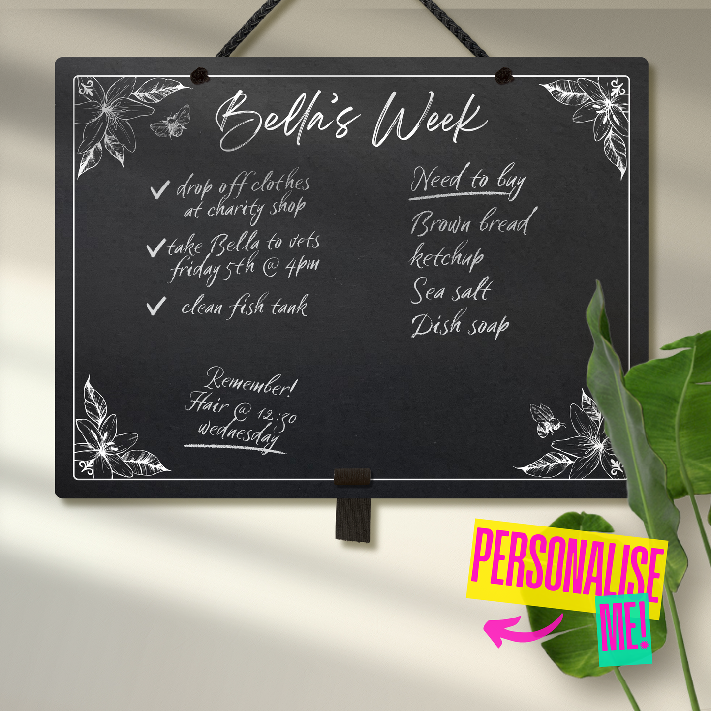 Large landscape chalkboard hanging with black rope and an adjustable pen holder at the bottom. Featuring sketchy floral illustrations framing a mostly empty space with 'Bella's Week' at the top in cursive font. A colourful sign reads 'Personalise me' whilst pointing towards the product.