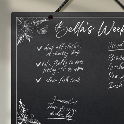 Close up of the large landscape chalkboard hanging with black rope, featuring sketchy floral illustrations framing a mostly empty space with 'Bella's Week' at the top in cursive font.