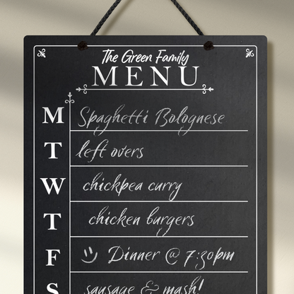 Close up of a large chalkboard hanging from black rope, featuring the days of the week printed down the left hand side with sections for each day and the heading 'The Green Family Menu' at the top. 