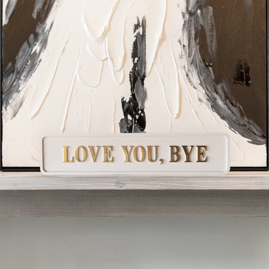 Cream and gold foiled street sign with "love you, bye" lettering on a shelf in front of abstract artwork