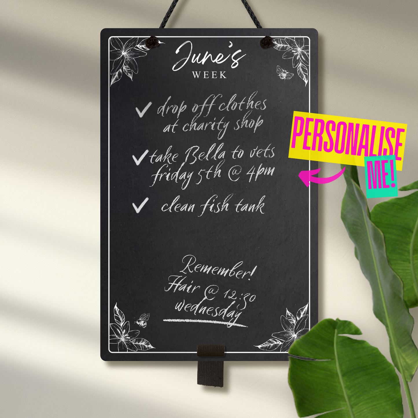 Medium portrait chalkboard hanging with black rope and an adjustable pen holder at the bottom. Featuring sketchy floral illustrations framing a mostly empty space with 'June's Week' at the top in cursive font. A colourful sign reads 'Personalise me' pointings towards the product.