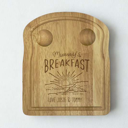 engraved wooden toast-shaped board with 2 carved pockets at the top for holding eggs, an engraved sunset design with 'mummy's breakfast' above and a personalised message at the bottom.