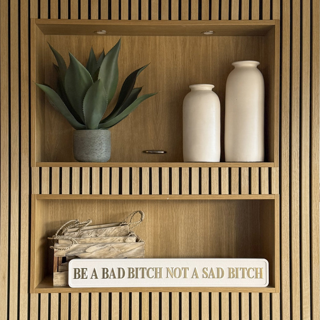 Wooden shelf with decorative items including a plant, vases, and a cream sign with gold text saying BE A BAD BITCH NOT A SAD BITCH.