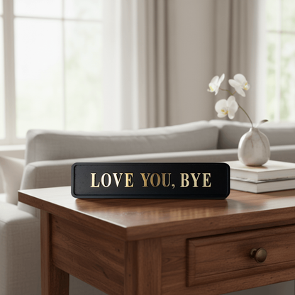 Black and gold foiled street sign with "love you, bye" lettering on a coffee table