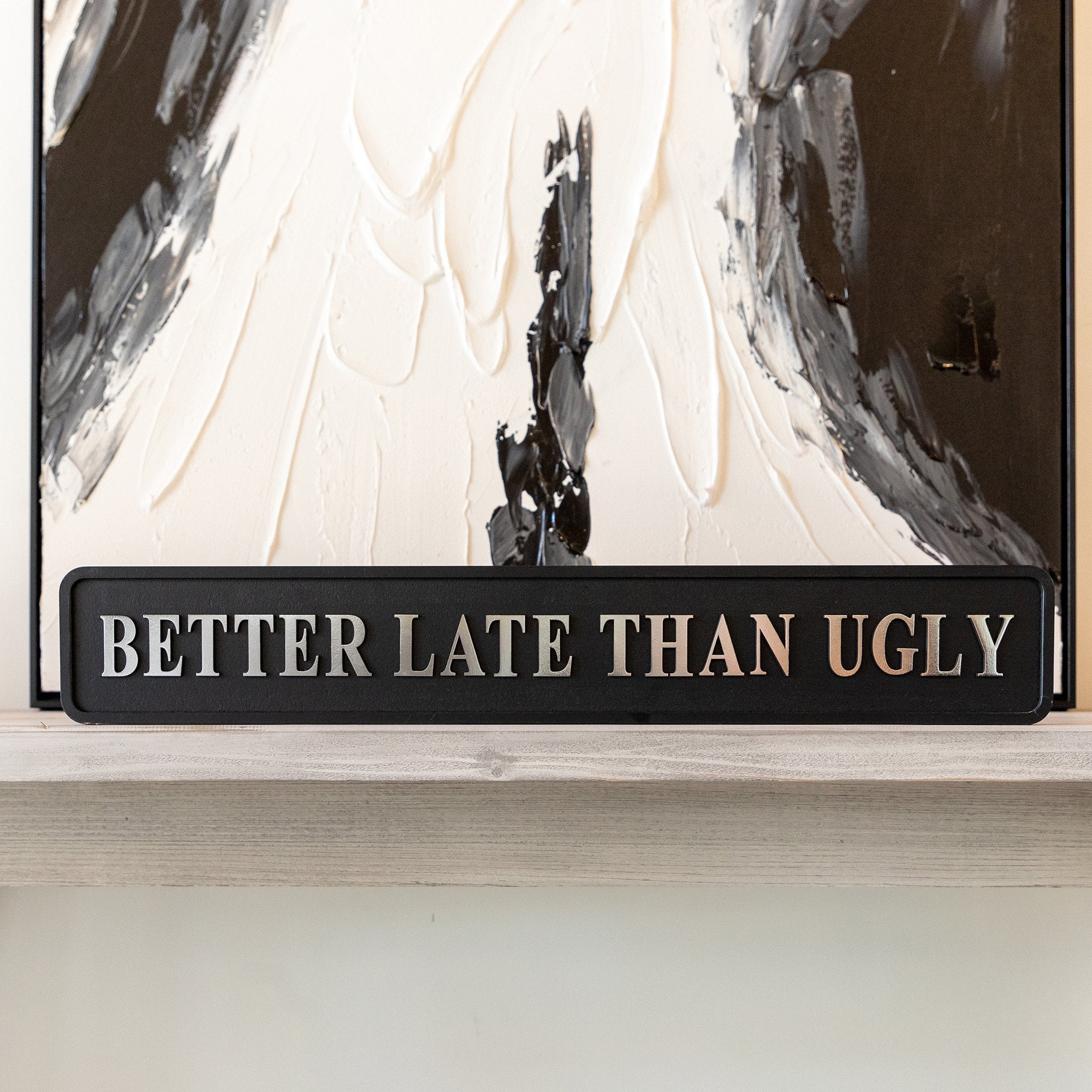 Decorative sign with 'Better Late Than Ugly' silver text in front of abstract art