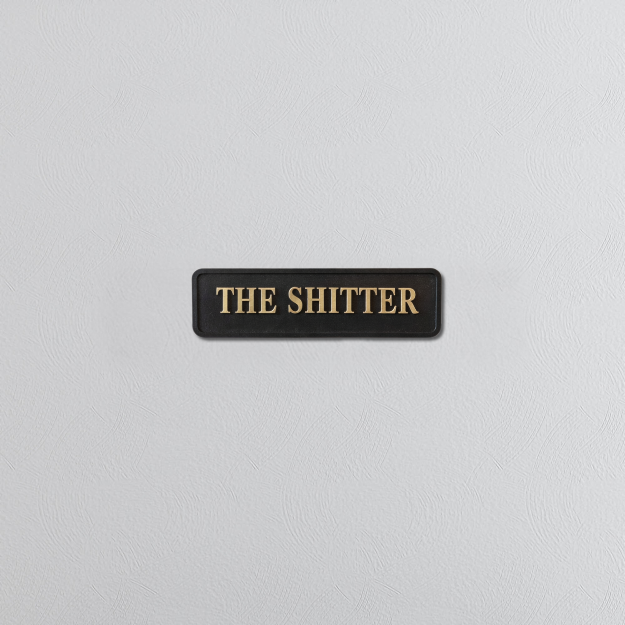 Black decorative sign with gold text 'THE SHITTER' on a grey background